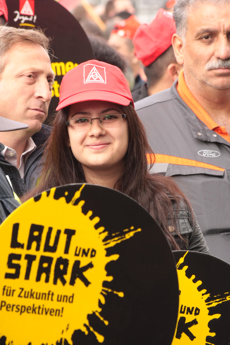 Demonstranten in Ford-Arbeitskluft und Demonstrantin mit IG-Metall-Mütze und Schild: »Laut und stark für Zukunft und Perspektiven!«.